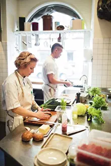 Cutting Prints: Female chef is cutting vegetables on board by colleague working at counter in restaurant kitchen