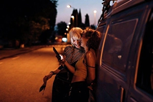 Road Trip Prints: Female couple laughing with phone at night, leaning on van