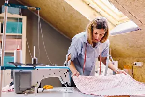 Focused Prints: Female tailor examining pink fabric while standing near table with sewing machine in attic workshop