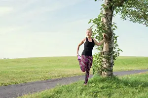 Runner Prints: Fit female athlete stretching leg near tree during training in meadow