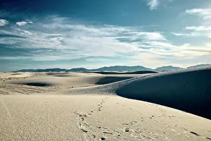 Arid Prints: Footprints trail across white sand dunes with distant mountains under a cloudy blue sky