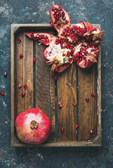 Ingredient Prints: Fresh ripe pomegranates cut in pieces in rustic wooden tray over dark blue plywood background