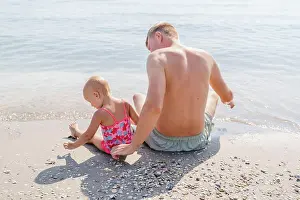 Coast Prints: Girl baby toddler with dad sit on the beach by the sea, back view