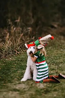 Gradeschooler Prints: Girl in elf hat kisses golden doodle dog in santa hat in grass