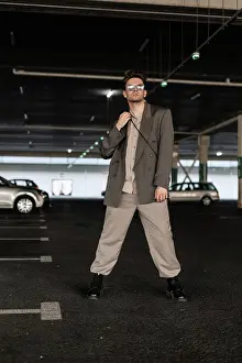 Stadt Prints: Gut aussehendes männliches Geschäftsmodell mit Sonnenbrille in modischem Outfit mit Stiefeln steht auf dem Parkplatz