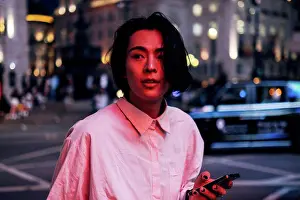 20 To 25 Years Prints: Handsome young man with long black hair holding mobile phone in city at night