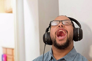 Disorder Prints: Happy African American man with Down Syndrome listening to music and singing with headphones at home