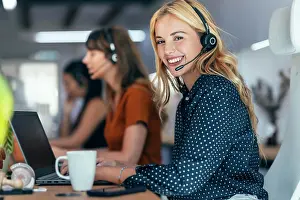 Conversation Prints: Happy blond businesswoman talking through headset sitting by colleagues at desk in office