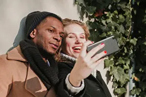 Multiethnic Prints: Happy multiethnic couple in warm clothes standing at bush and taking selfie