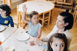 Stockholm Prints: High angle view of girl with arm raised sitting amidst teacher and friends at table in classroom