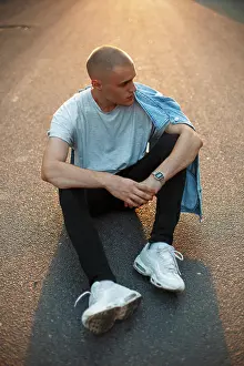Stadt Prints: Hübscher junger Mann in grauem T-Shirt und modischer Jeans sitzt bei Sonnenuntergang auf der asphaltierten Straße ***