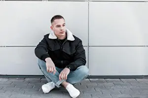 Stadt Prints: Hübscher junger Mann in stilvollen Jeans in einer trendigen Jacke in weißen Turnschuhen mit einer Frisur entspannt