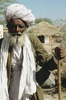 Staring Prints: India, Rajasthan, Senior local man leaning on cane, village houses in background