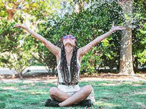 Trendy Prints: Joyful young female with dreadlocks sitting on ground legs crossed throwing colorful confetti and looking up