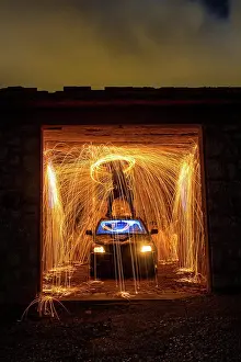 Anonymous Prints: Long exposure of silhouette of human with burning circle standing on roof of automobile in shed between fireworks