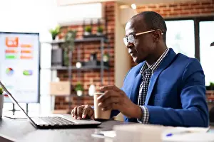 Male employee holding paper cup and working on his personal computer in startup office