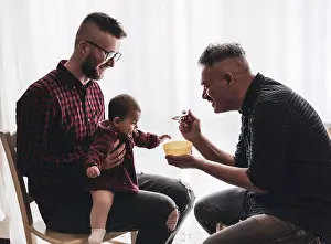 Dads Prints: Male gay couple with adopted baby girl at home - Two handsome dads feed the baby girl on kitchen - Male babysitters