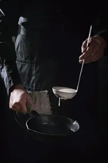Back Ground Prints: Man chef in black apron pouring dough from ladle for cooking pancakes in cast-iron pan