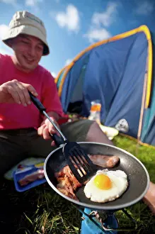 Tent Prints: Man cooking fried breakfast of bacon and egg beside tent; Lulworth, Dorset, UK