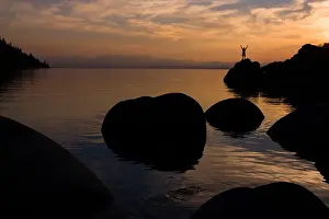 Hike Prints: A man standing on a rock on the shore of Lake Tahoe in California at sunset with reflecting water