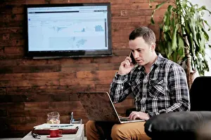 Midadult Men Prints: Man using a mobile phone using laptop sitting on sofa in a small office