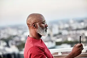 Bald Prints: Mature bald man with white beard using smart phone for photographing sunset from rooftop