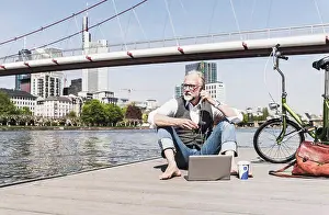 Best Prints: Mature man with laptop, earbuds and bicycle sitting at the riverside in the city