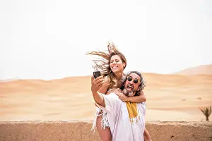 Balcony Prints: Middle-aged man with woman piggyback taking selfie expressively on terrace against sandy desert