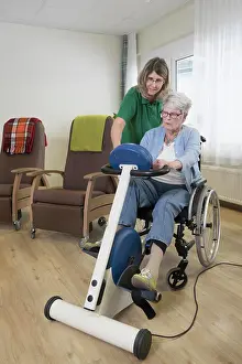 Disability Prints: Nurse with senior woman on wheelchair exercising on exercise bike in rest home