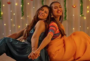 Occasion Prints: Portrait two cheerful young women sitting back-to-back during Diwali celebration