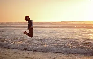 30 To 35 Years Prints: Portrait of a laughing woman leaping into the air while standing at the edge of the sea on a beach