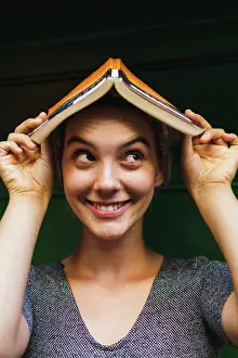 Smart Prints: Portrait of young playful woman smiling away and holding opened book on head