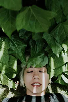 Out Door Prints: Portrait of young smiling girl with blonde hair resting with eyes closed while lying on green leaves in sunlight