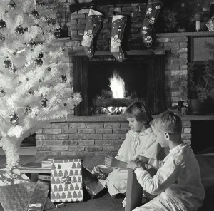 Opening Prints: Side profile of a girl and her brother unwrapping Christmas presents near a fireplace
