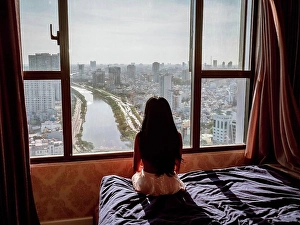 City View Prints: Rear view of an Asian woman sitting on a bed and looking through the window to the city of Ho Chi Minh