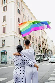 Gender Prints: Rear view of young lesbian couple standing on a street, waving rainbow flag