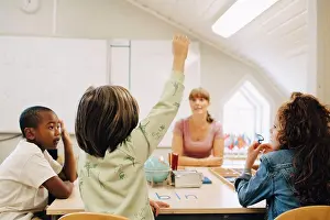 Communications Prints: Schoolboy raising hand while answering teacher in classroom at school