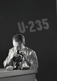 Scientist Prints: Scientist looking through microscope with U-235 (Uranium Isotope) being projected on the wall