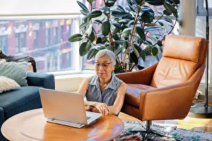 Asian And Indian Ethnicities Prints: Senior Asian Woman sitting and using laptop at home in living room