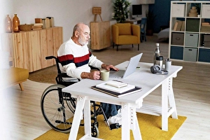 Handicapped Prints: Senior businessman on wheelchair with coffee mug using laptop at home office