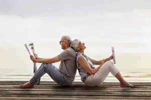 Husband Prints: Senior couple sitting back to back on wooden dock near lake and reading newspaper