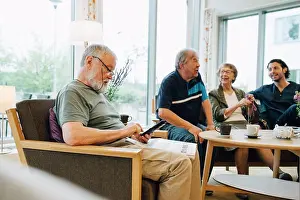 Using Phone Prints: Senior man using smart phone while sitting with newspaper on armchair by friends and caretaker at elderly nursing home