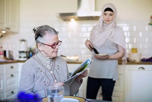 Newspaper Prints: Senior woman reading newspaper with female home caregiver preparing food in kitchen