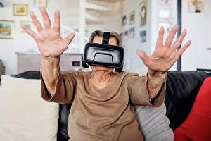 Top Section Prints: Senior woman wearing virtual reality simulator gesturing while siting on sofa in living room at home