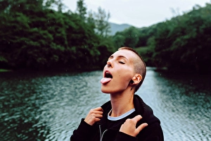 Tongue Out Prints: Short haired female with piercing wearing black hoodie looking up while catching raindrops with tongue near green