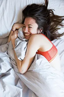 Nose Ring Prints: From above shot of young laughing brunette with nose ring wearing red lingerie and cuddling in bed