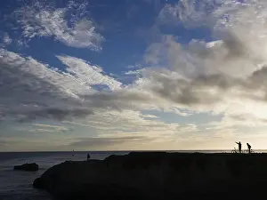 Small Group Prints: Silhouette of people with bicycles on rock at water with blue sky and clouds
