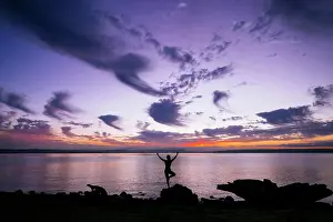 Meditation Prints: Silhouette of a woman performing a yoga pose on a rock by the waterside at sunset