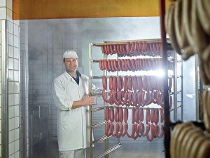 Manual Worker Prints: Smiling butcher standing by arranged sausages in smokehouse