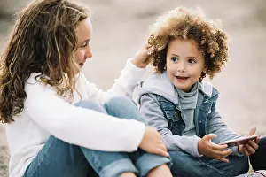 Gadget Prints: Smiling casual curly child resting on ground with crossed legs holding mobile phone while content girl sitting clasping